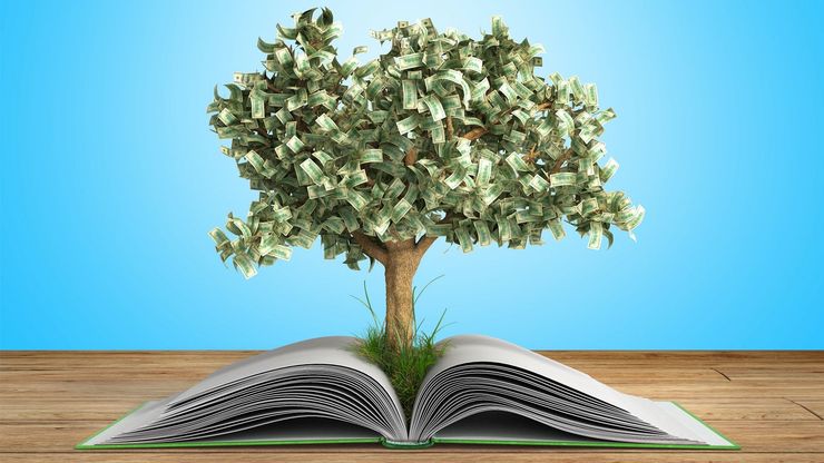 An open book on a wooden surface sprouting a tree made from money against a blue background. 